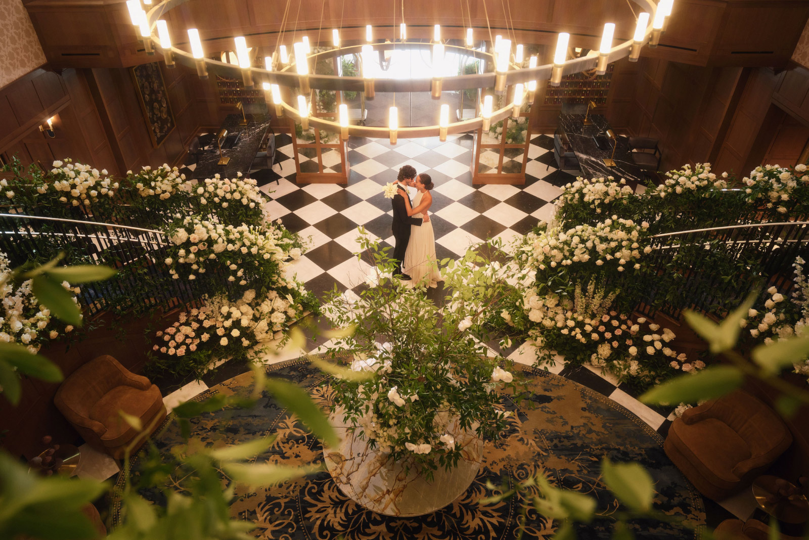 A romantic scene of a couple sharing a dance in an elegantly decorated hotel lobby, framed by lavish floral arrangements and illuminated by a grand circular chandelier overhead.