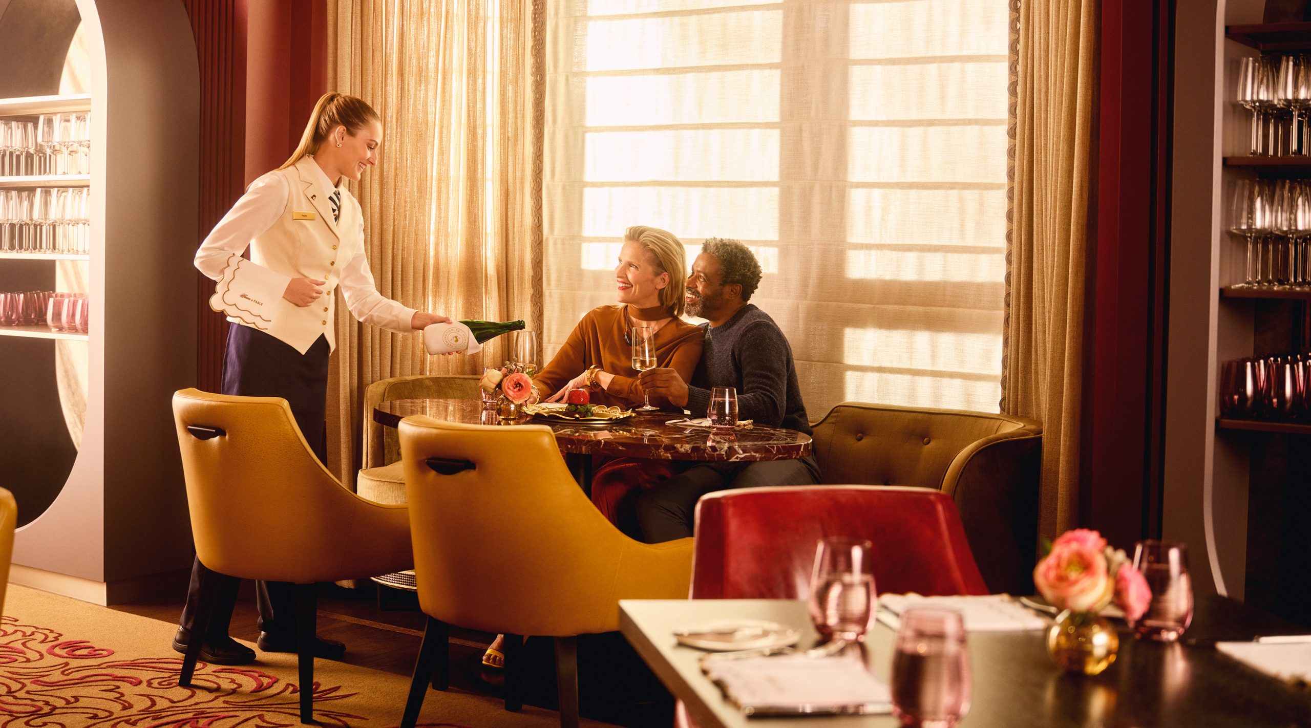 Woman and man sitting at a comfortable table at Fawn & Fable restaurant at Nemacolin resort as a female server pours a glass of wine for them