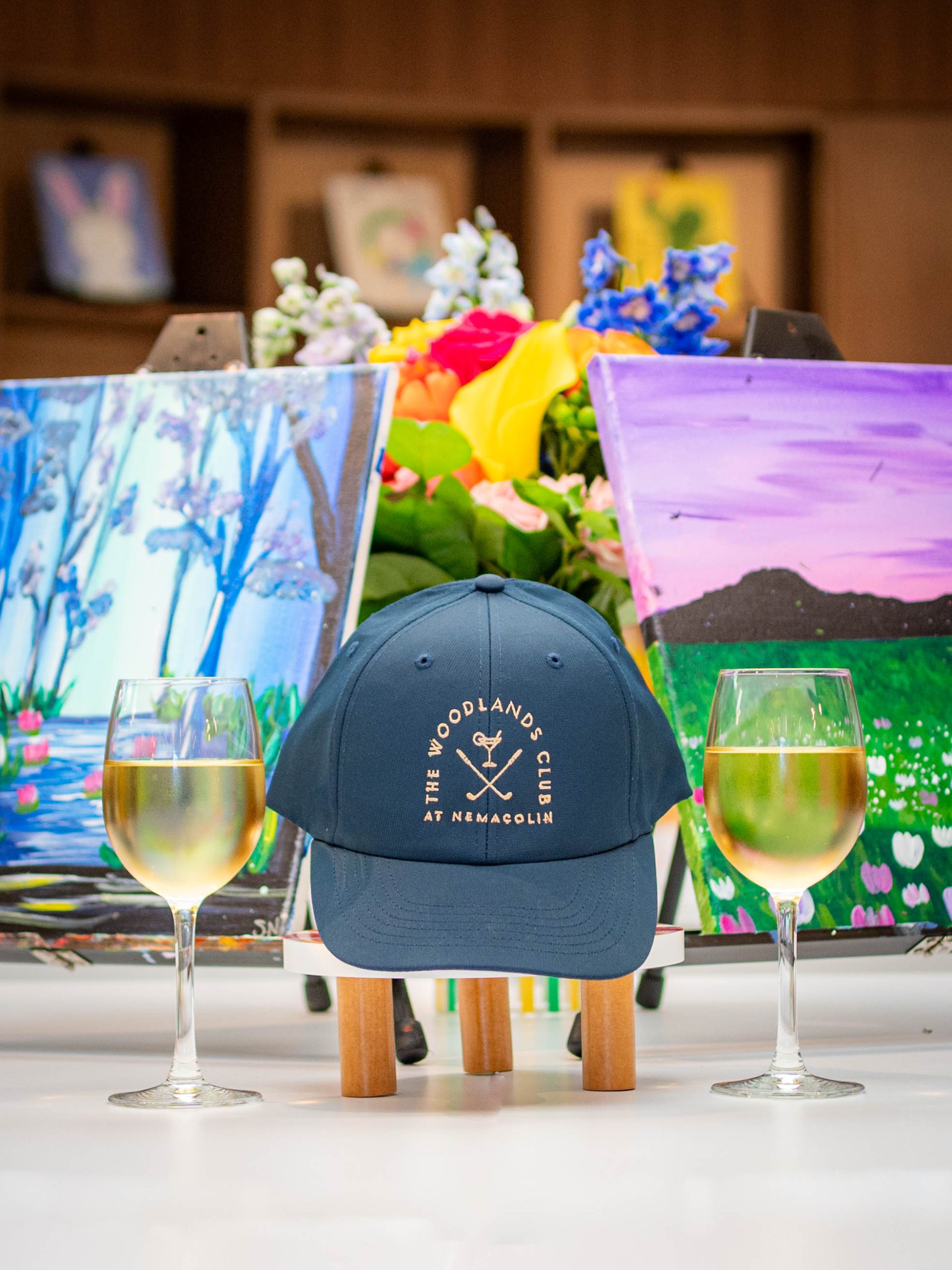 A blue hat with the logo of The Woodlands Club at Nemacolin is surrounded by two glasses of white wine, two canvas paintings, and a bouquet of flowers.